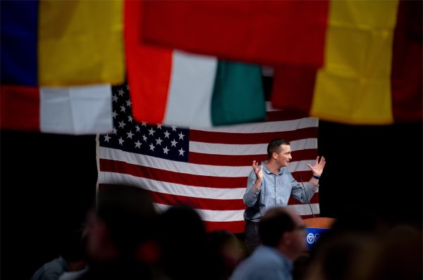 Nicholas Maodush-Pitzer, sixth-grade teacher at Blandford Elementary, stands at podium with arms raised; American flag in background, other flags hanging from ceiling