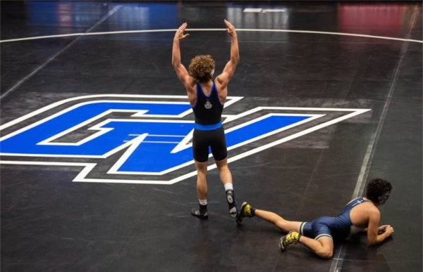A wrestler holds arms up in triumph while another wrestler lies on the floor. The mat has GV in the center.