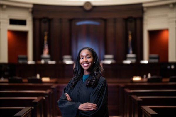 A person wearing a justice's robe smiles with arms folded in a courtroom.