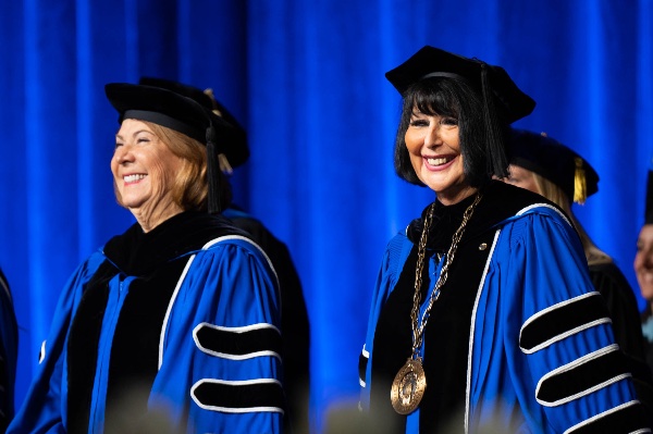 Two people in academic regalia stand on stage smiling. 