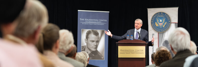 Jon Meacham stands at a podium and gestures to the crowd in the middle of a packed auditorium. The backs of the heads of the audience are blurred in the foreground, Meacham is in sharp focus in the back.
