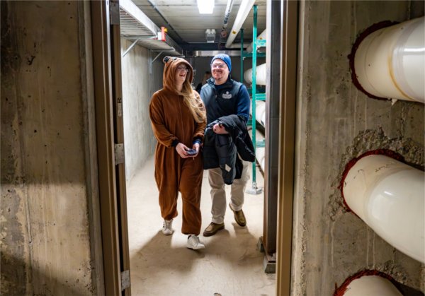 A young woman in a groundhog onesie walks with a man through the basement of Lake Ontario Hall. 