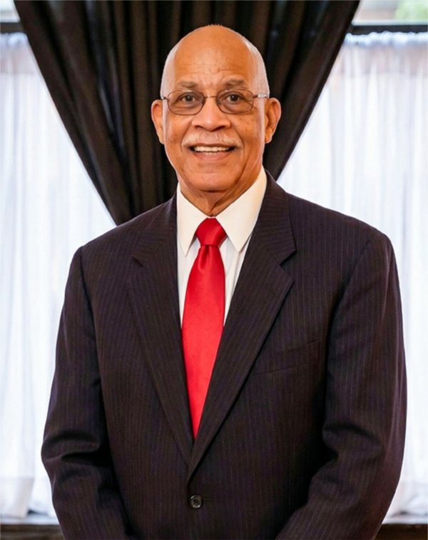 Don Williams in dark suit jacket, white shirt, red tie in front of window with black drapes pulled in.