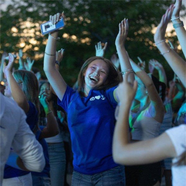 A smiling person dancing in a crowd with their hands held up.