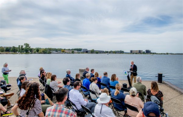 A man speaks at a podium in front of an audience with Muskegon Lake in the background.