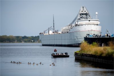 A man fishes from a boat on Muskegon Lake with a large freight docked behind him.