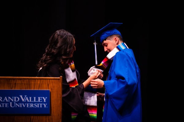 woman pins something to a stole worn by a student in a cap and gown on stage