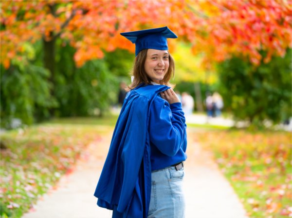 A person wearing a cap and with a graduation gown slung over their shoulder smiles.
