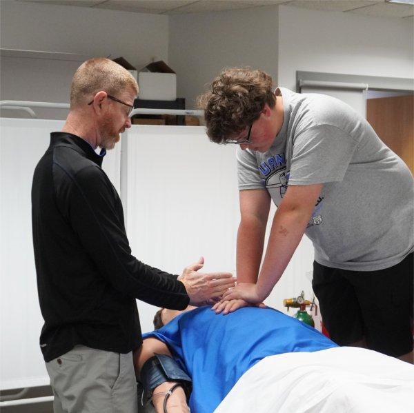 A student practices chest compressions on a medical training mannequin while an instructor observes and provides guidance in a classroom lab setting