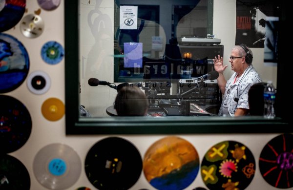 A person is seen through glass talking in front of radio studio equipment in a radio studio. Vinyl records and CDs are taped to the wall.