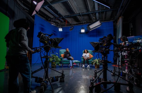 Two people smile while sitting in chairs in a simulated television studio. A person works a camera in the foreground.