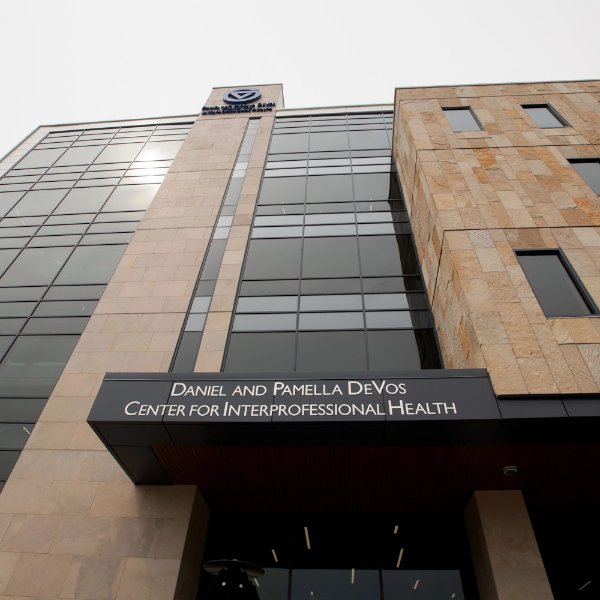 The exterior of a multi-story building shown from ground level. The entrance has the words, "Daniel and Pamella DeVos Center for Interprofessional Health."