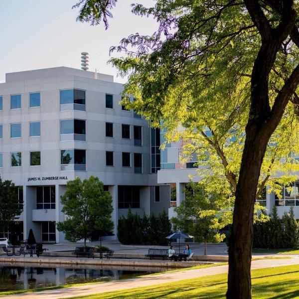 James H. Zumberge Hall is in the background, along with Zumberge Pond. A tree is in the foreground. People are sitting at a table with an umbrella.