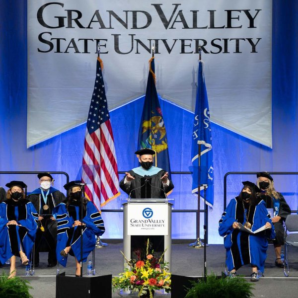 speaker at podium gestures with open arms toward audiences; others in academic regalia on the platform; GVSU banner hangs down