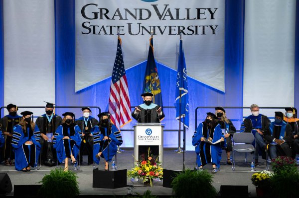 man in academic regalia gestures toward audience, others on platform also in regalia; GVSU banner hangs down