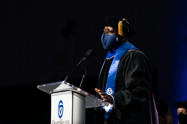 man at podium in academic regalia and wearing face mask