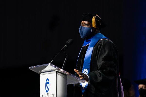 man at podium in academic regalia and wearing face mask