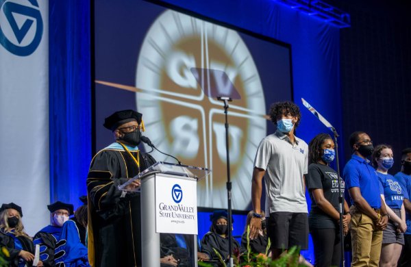 B. Donta Truss, vice president for Enrollment Development and Educational Outreach, on platform in academic regalia and line of students in front of screen with GVSU bursting out