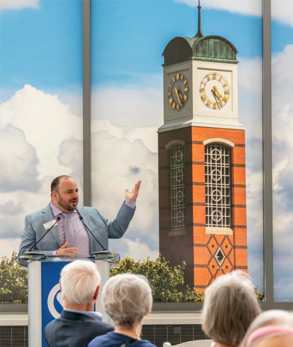 Jon Lehrer stands at podium during a meeting when he was introduced as the carilloneur, image of Cook Carillon on backdrop behind him