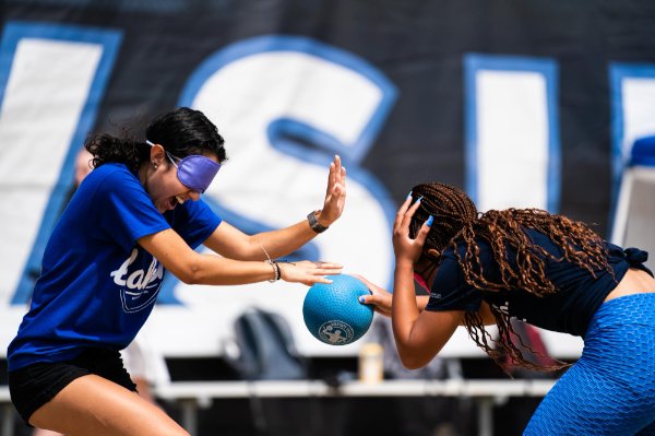   Two people wearing blindfolds hold their arms out, one holds a ball in their hand.