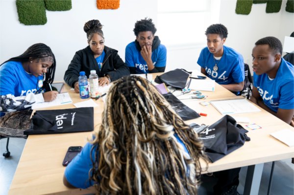 A group of students in blue shirts interview a young woman who sits at the other end of their table. 