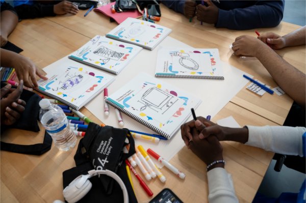 A group lay out their five notebooks and compare prototype designs that they have drawn. 