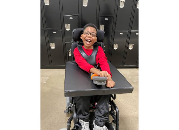 young child in wheelchair with arm on device in front to control it; kid is wearing glasses and a red shirt