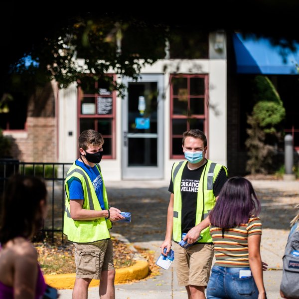 Two male GVSU students in yellow vests give another GVSU student a reusable face covering on the Allendale Campus.