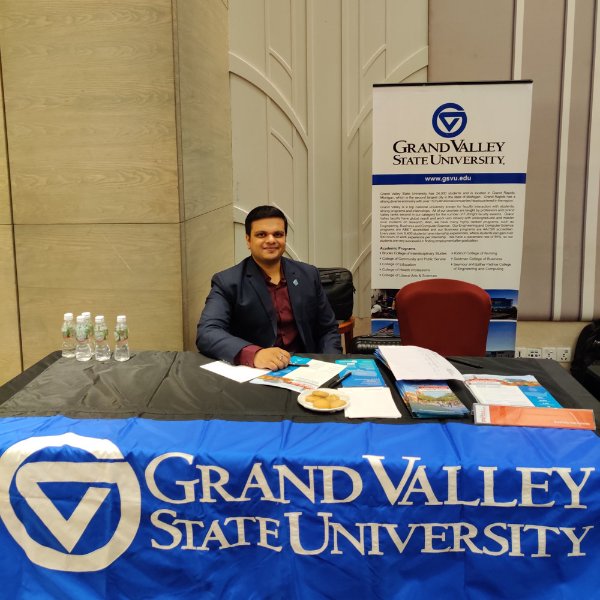 Kirthi Kondapalli sits behind a table with a GVSU blue banner in front, and recruitment materials, bottles of water