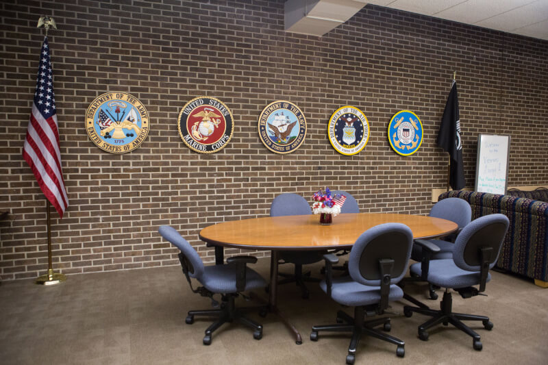 A photograph of the Veterans Lounge on campus in the Kirkhof Center. The logos of each of the armed forces are displayed on the wall, with the American Flag to the left of the image. 