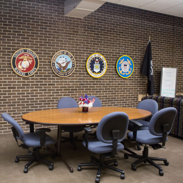 A photograph of the Veterans Lounge on campus in the Kirkhof Center. The logos of each of the armed forces are displayed on the wall, with the American Flag to the left of the image. 