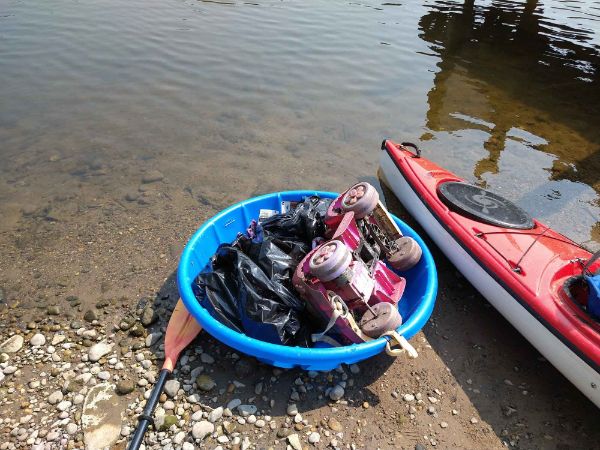 Along a rocky river bank, a kayak is next to a small plastic pool containing a black plastic garbage bag and a toy motorized car. An oar sits next to the tub. 
