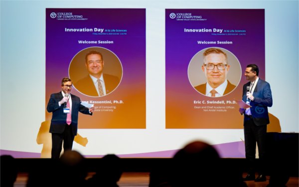 Two speakers stand on a stage holding microphones during the Innovation Day welcome session, with large projected slides behind them displaying headshots and titles of Marouane Kessentini and Eric Swindell.