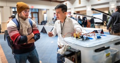 A presenter speaks animatedly to an attendee while demonstrating a device with visible wiring and electronic components at a display table.