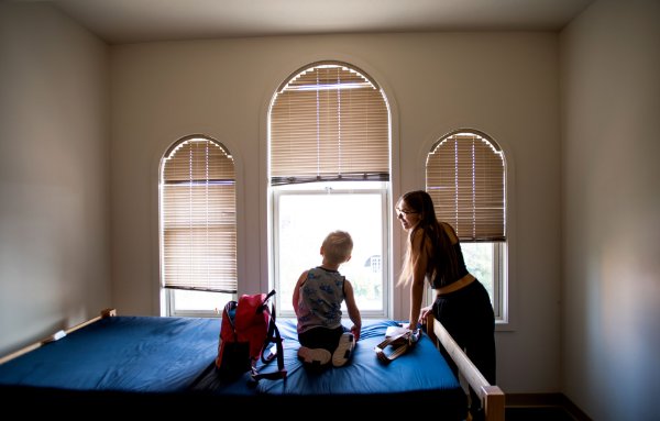 An adult, standing next to a bed, looks out the window with a child who is sitting on the bed.