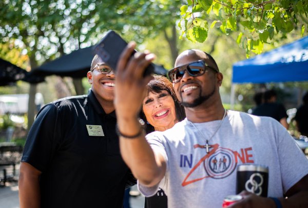 Three people smile while one person takes a selfie with a phone.