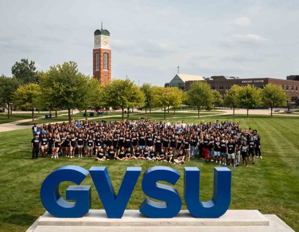 A large group of people pose for a photo behind the large GVSU letters. The carillon is in the background.