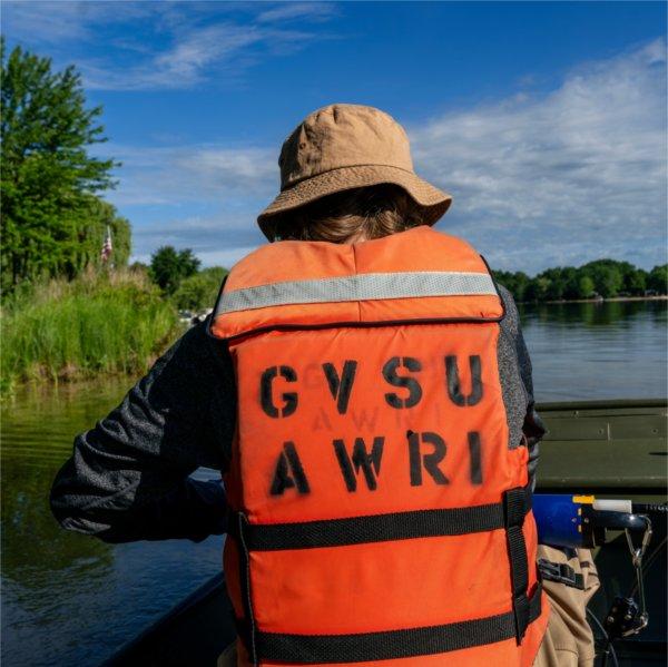 A researcher wearing an orange Grand Valley State University Annis Water Resources Institute life jacket and tan bucket hat sits in a small boat on a calm lake, facing the shoreline.