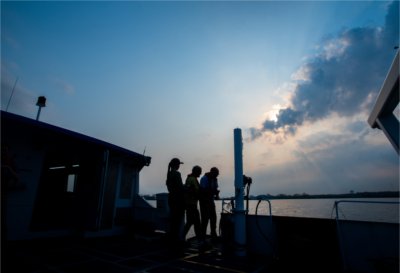 Researchers stand on the deck of the Grand Valley State University Annis Water Resources Institute's W.G. Jackson research vessel as sun rays break through clouds over the water.