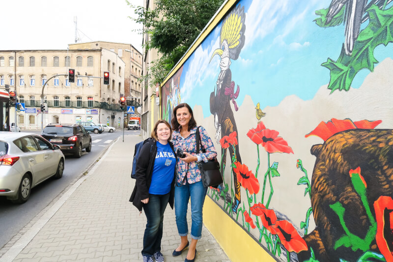 two women stand on sidewalk in front of mural