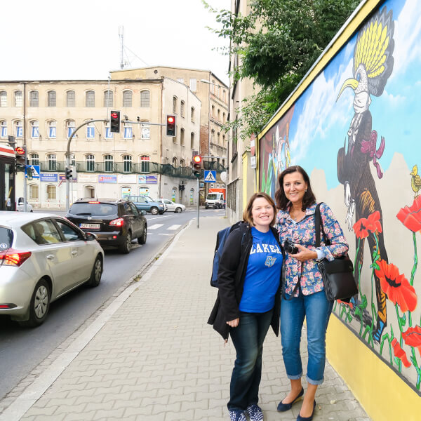 two women stand on sidewalk in front of mural