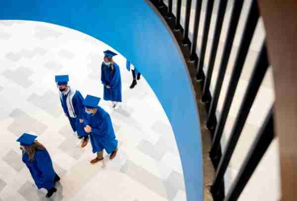 Students wearing caps and gowns walk under a staircase in the fieldhouse