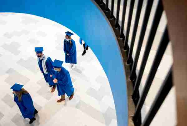 Students wearing caps and gowns walk under a staircase in the fieldhouse