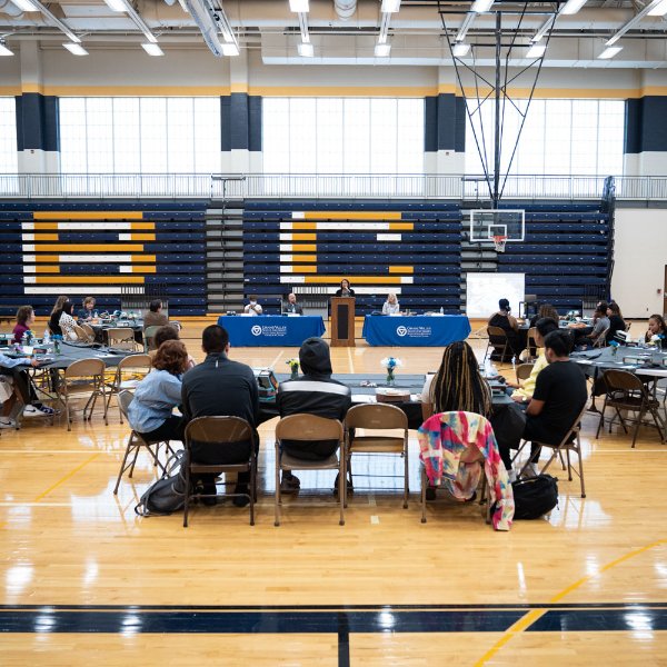 tables in a gymnasium with people seated around them; head table has blue drapes with GVSU logo. Bleachers in background are blue with large gold B and C showing