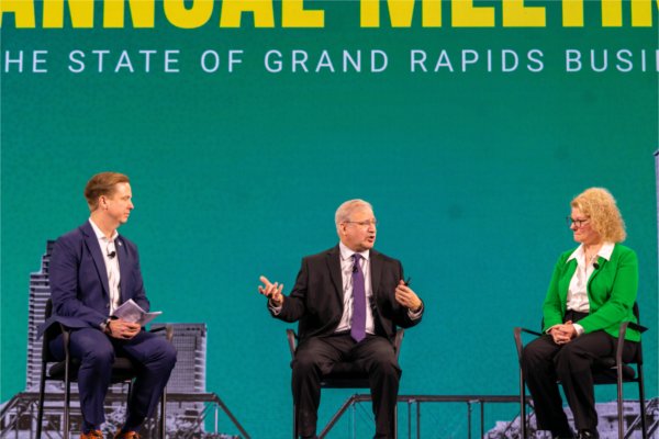 Three panelists sit on stage at Grand Rapids Chamber's Annual Meeting, discussing the state of Grand Rapids business in front of a large green backdrop.