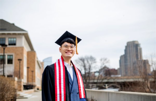 A person wearing a cap and gown smiles while looking off into the distance. Buildings are in the background.