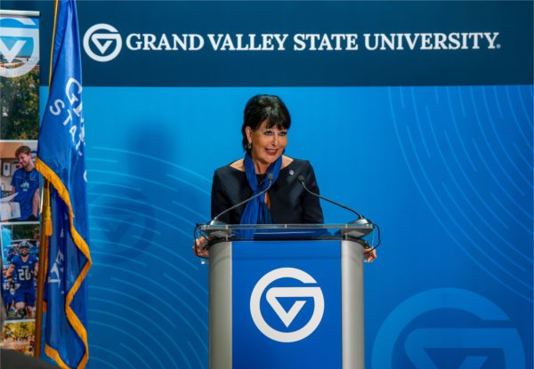 A person smiles while standing at a podium. The words Grand Valley State University are on a banner in the background. A flag is also in the background.