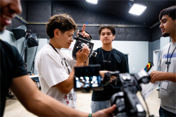 A young man looks through the viewfinder of a camcorder as two other people watch.