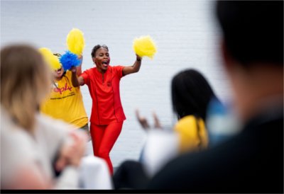 A teacher wearing all red holds up yellow pompoms and cheers on her students presenting. 