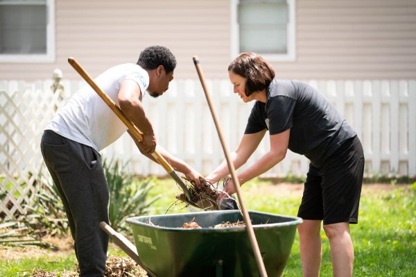 One person holds a shovel near a wheelbarrow while another person helps with the contents of the shovel.